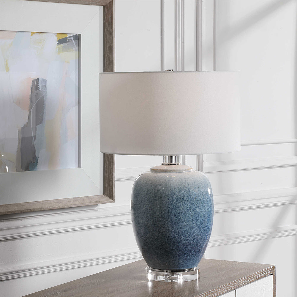 Front facing image of lamp. Ombre blue base that fades to white with a crisp white drum shade. The lamp sits on a crystal disc, with nickel accents. Lamp is sitting on a timber console with a piece of abstract art behind.