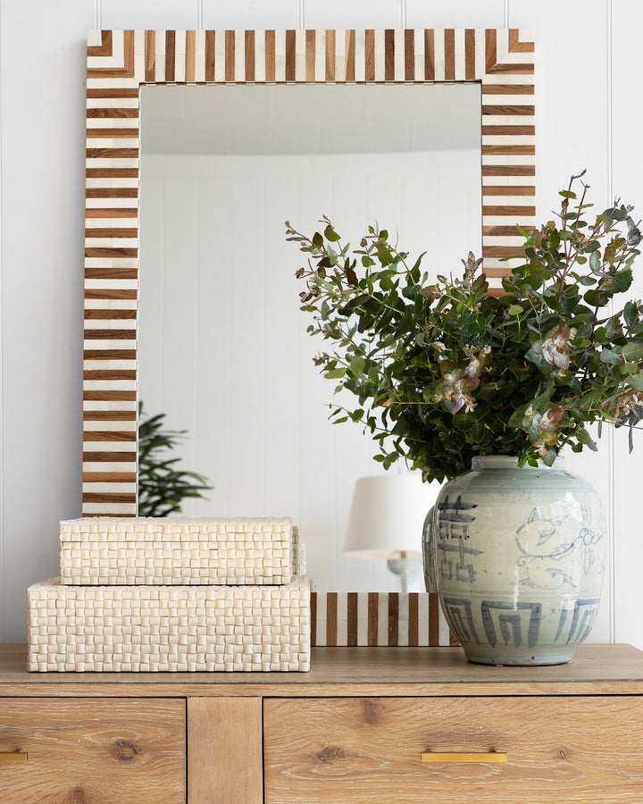Wooden dresser with decorative items including a striped mirror, boxes, and a vase with flowers.