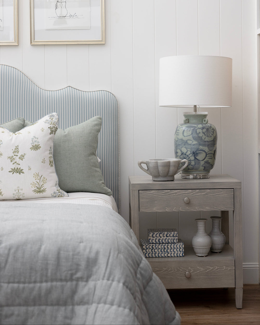 Light coloured timber bedside table with pale blue lamp and blue striped bedhead