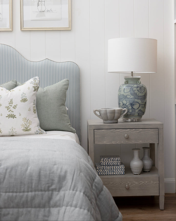 Light coloured timber bedside table with pale blue lamp and blue striped bedhead
