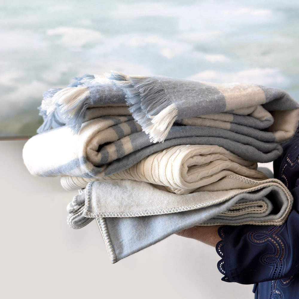 A stack of folded woven blankets in soft gray, cream, and beige tones, held by a person whose arm and dark blue sleeve are visible against a blurred outdoor water background.