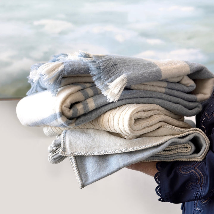 A stack of folded woven blankets in soft gray, cream, and beige tones, held by a person whose arm and dark blue sleeve are visible against a blurred outdoor water background.