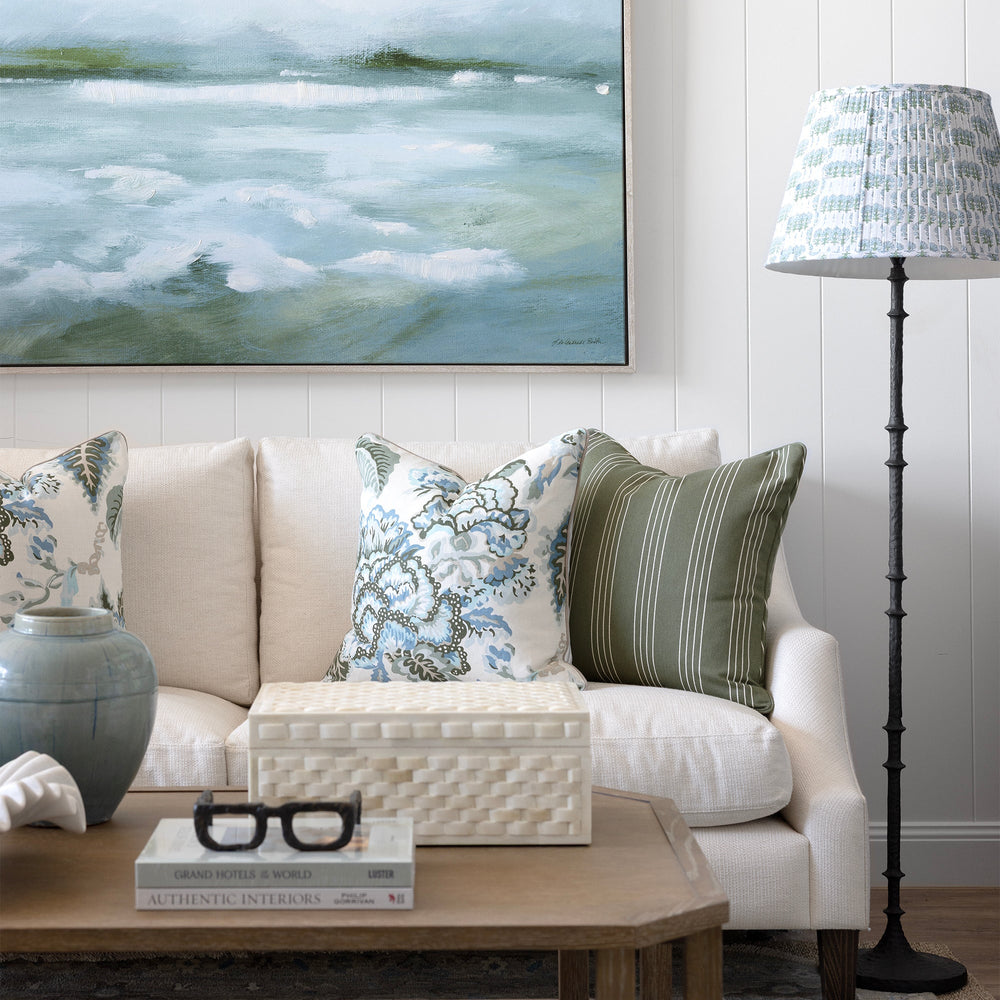 A large framed landscape painting of misty waves hangs on a white paneled wall above a cream sofa with patterned throw pillows. A wooden coffee table, a blue-gray ceramic vase, and a black floor lamp with a patterned shade are visible in the styled room.