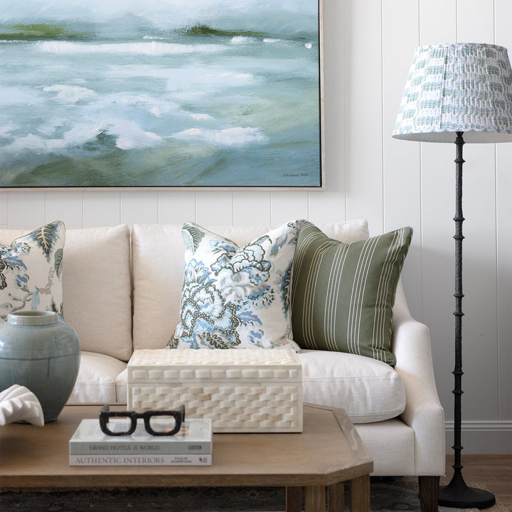 A large framed landscape painting of misty waves hangs on a white paneled wall above a cream sofa with patterned throw pillows. A wooden coffee table, a blue-gray ceramic vase, and a black floor lamp with a patterned shade are visible in the styled room.