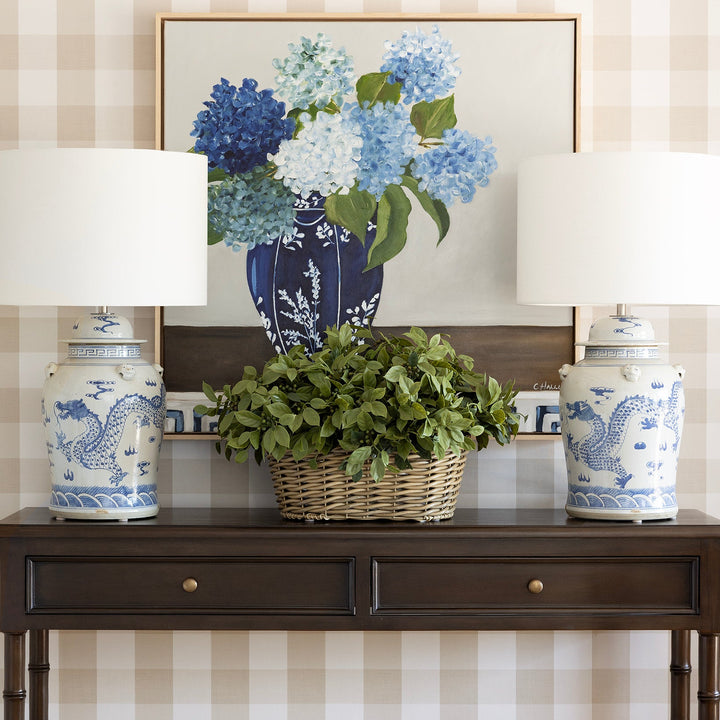 A dark brown wooden console table styled in a home entryway, flanked by two white table lamps with blue dragon-painted ceramic bases. A wicker basket filled with green foliage sits in the center, and a framed floral print hangs on the beige-and-white checkered wallpapered wall behind it.