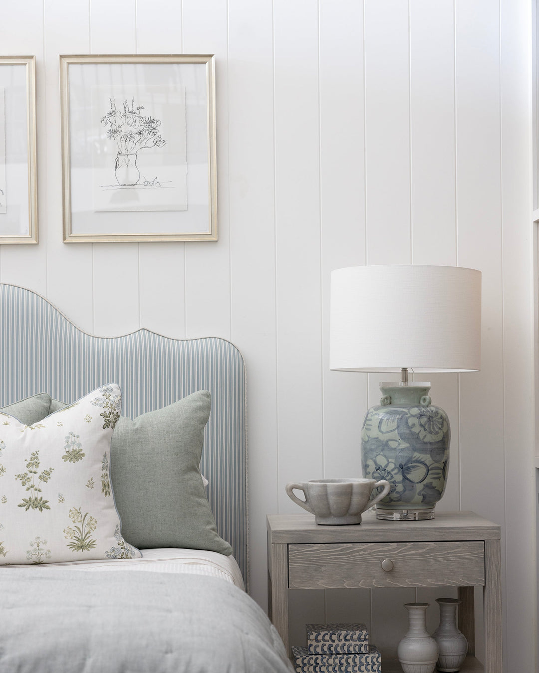 A styled bedroom corner featuring a light gray ribbed headboard, gray bedding, a gray nightstand with a blue and white patterned ceramic table lamp, and white paneled walls with framed botanical prints.