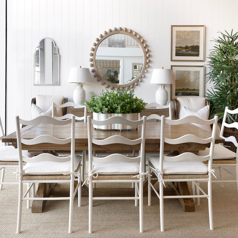 A dining room scene featuring a rustic wooden dining table surrounded by six white ladder-back chairs with white upholstered seats. The room is styled with a large round decorative mirror, white table lamps, framed art, potted green plants, and a white sideboard on a light beige rug.