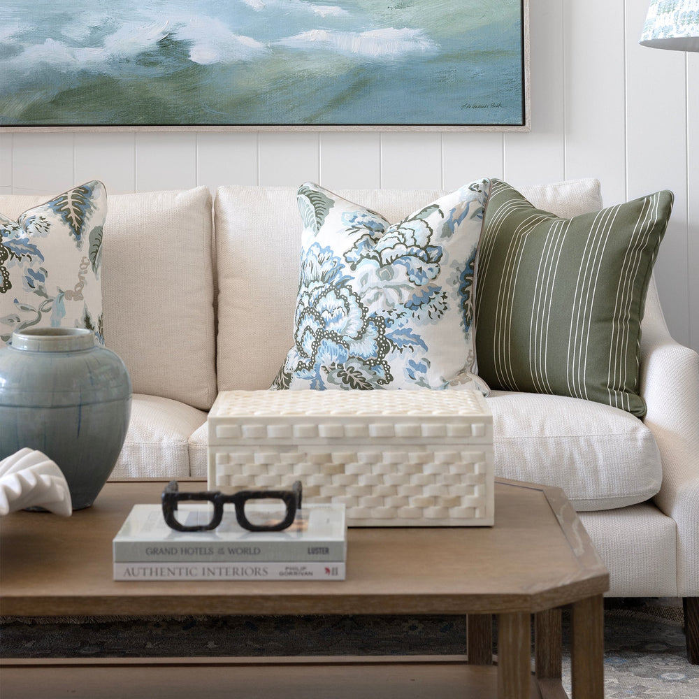 A large framed landscape painting of misty waves hangs on a white paneled wall above a cream sofa with patterned throw pillows. A wooden coffee table, a blue-gray ceramic vase, and a black floor lamp with a patterned shade are visible in the styled room.
