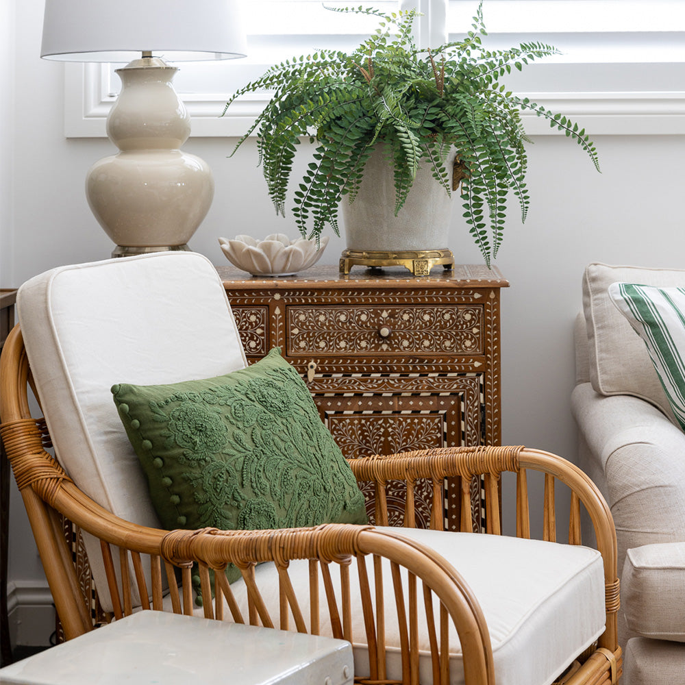 Cape Cod Rattan Armchair styled with a rich green embroidered cushion. The chair is sitting in front of a teak wood and bone inlay cabinet, styled with a faux fern arrangement a small marble bowl and a large cream lamp. 