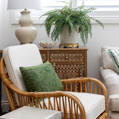Cape Cod Rattan Armchair styled with a rich green embroidered cushion. The chair is sitting in front of a teak wood and bone inlay cabinet, styled with a faux fern arrangement a small marble bowl and a large cream lamp. 