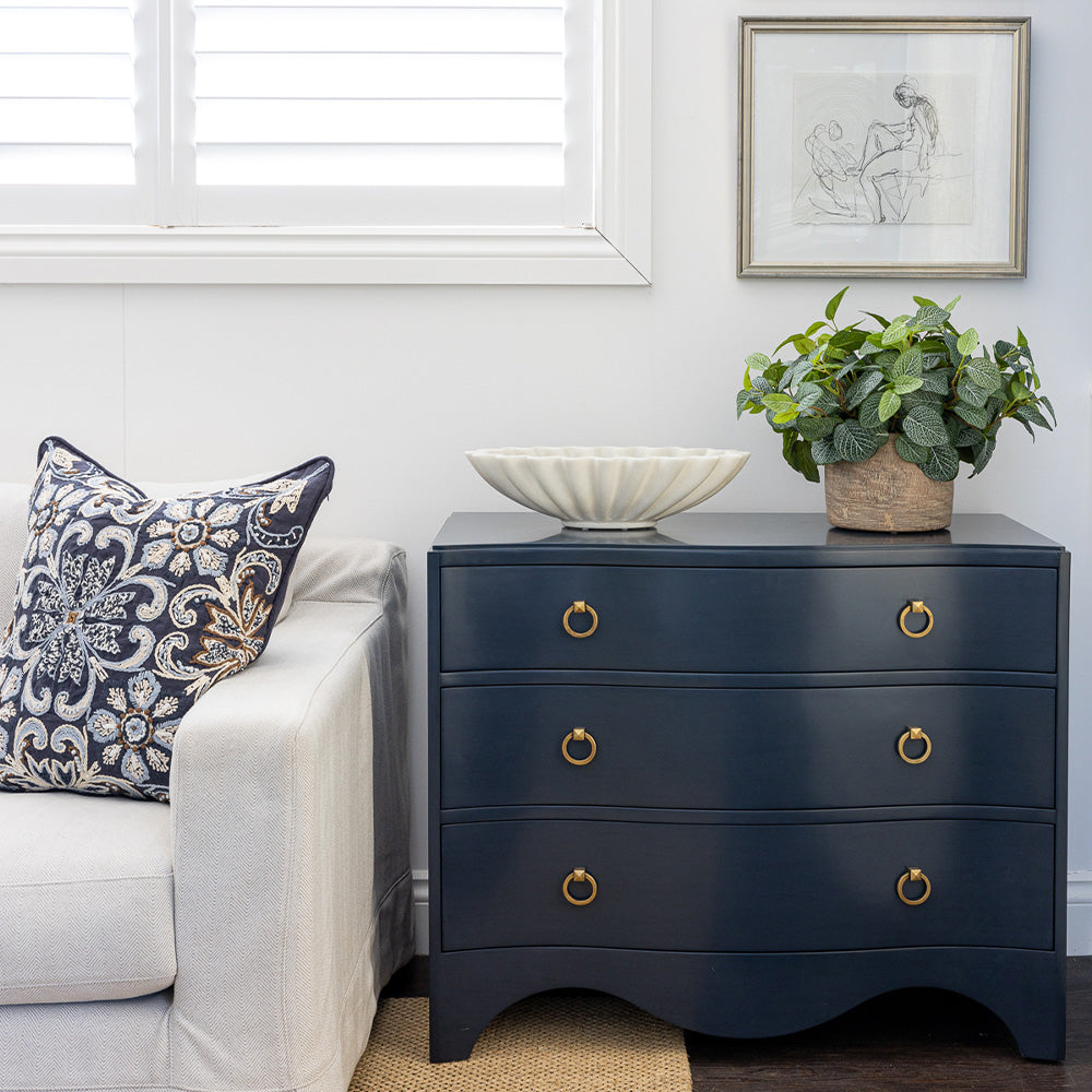Image of Navy Claire Chest styled next to a neutral sofa with a navy embroidered cushion. The chest is styled  with an oval ruffled marble bowl and faux fittonia greenery on top, figure study artwork hanging on wall above.
