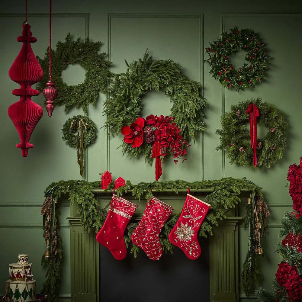 Decorative Christmas mantel with wreaths, stockings, and red ornaments against a green wall.
