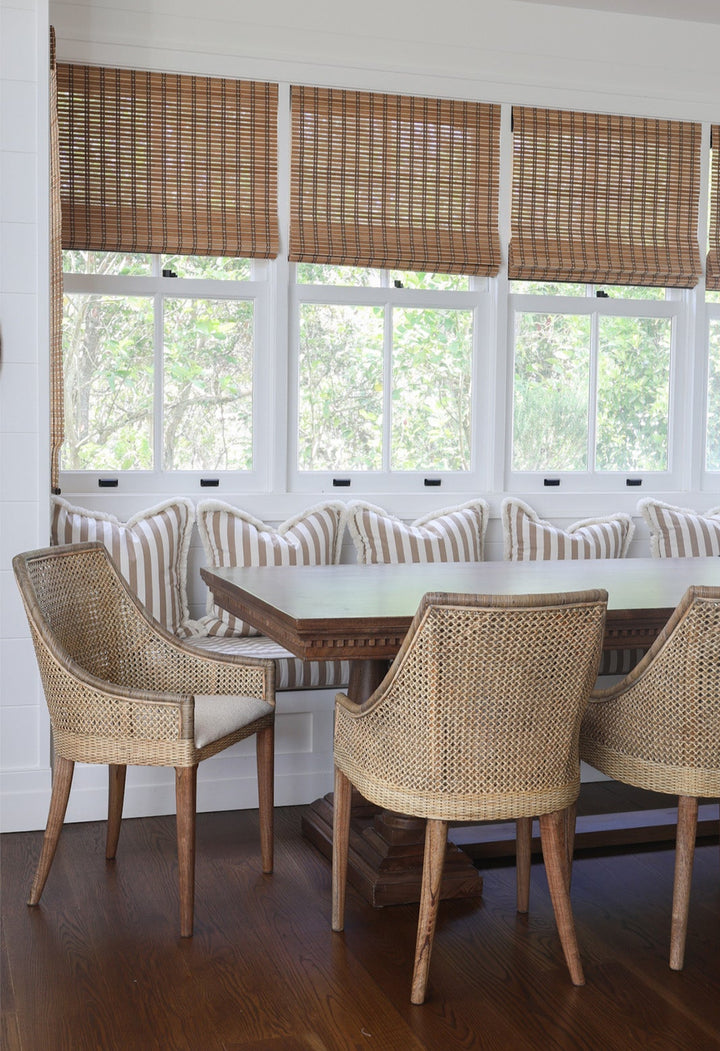 Dining room with rattan dining chairs, a long rectangular dark mango wood table, and striped bench against a window with woven blinds.