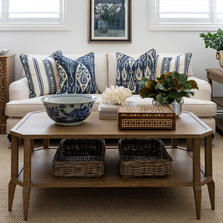 Image of Eliza coffee table styled in front of sofa. On top styled with a blue & white bowl, a teak and bone inlay box, a stack of three books topped with decorative coral, and a faux magnolia leaf arrangement. On the lower shelf are two wicker basket trays.