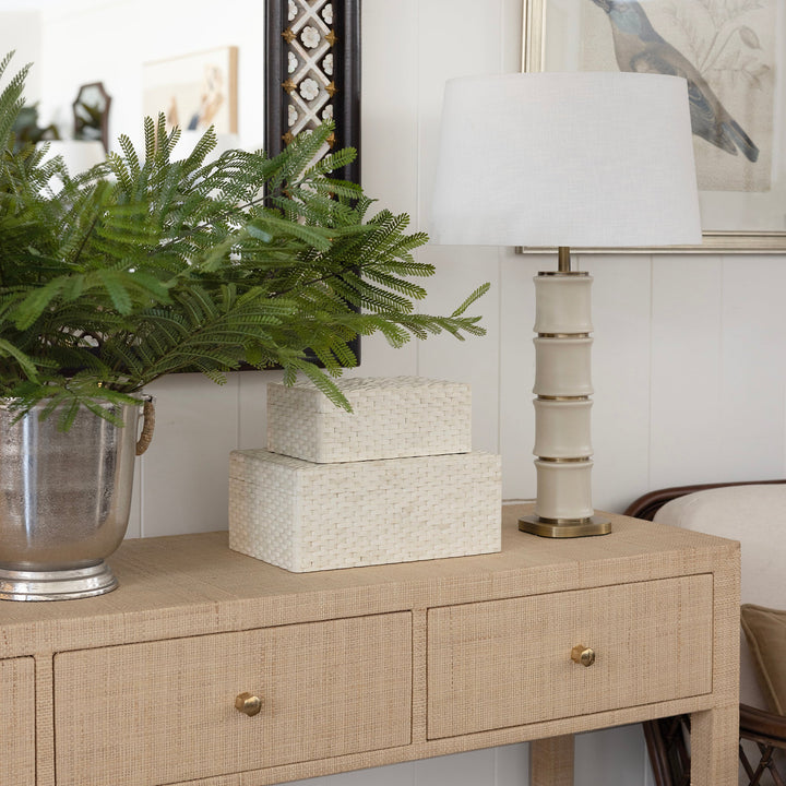 A raffia woven console table in a styled room setting, decorated with a green fern in a silver pot, two stacked white textured boxes, and a cream-colored table lamp with a white shade.