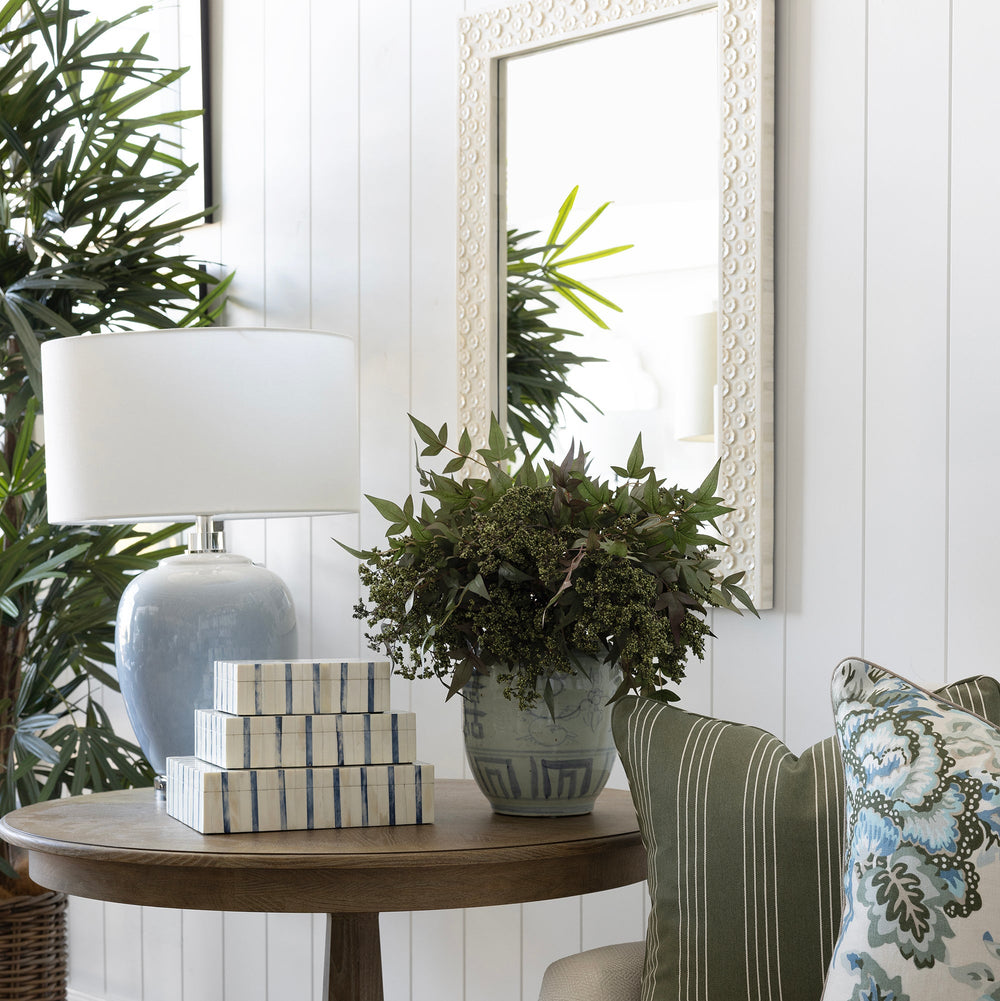 A styled interior corner featuring a round wooden side table with a white lamp, stacked decorative boxes, and a potted green plant, next to a cream sofa with green and floral throw pillows.