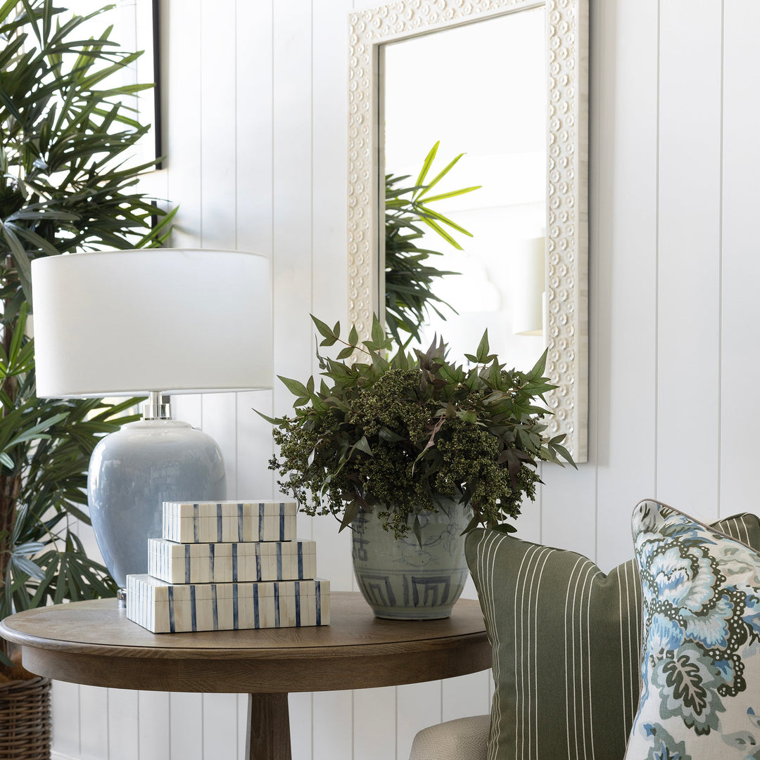 A styled interior corner featuring a round wooden side table with a white lamp, stacked decorative boxes, and a potted green plant, next to a cream sofa with green and floral throw pillows.