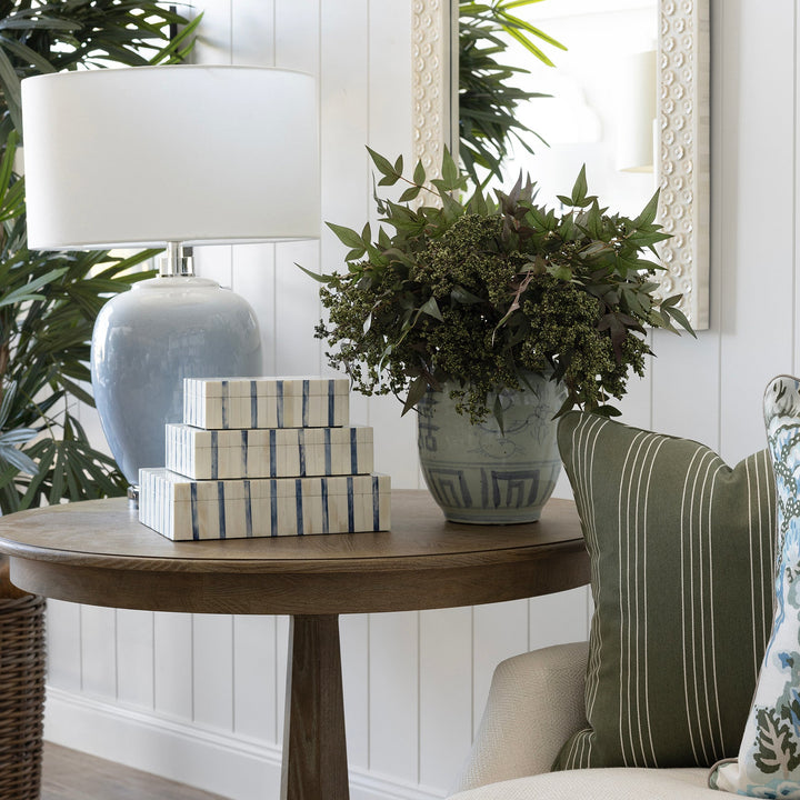 A styled interior corner featuring a round wooden side table with a white lamp, stacked decorative boxes, and a potted green plant, next to a cream sofa with green and floral throw pillows.