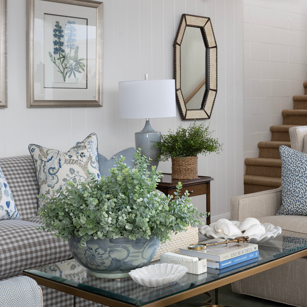 A styled living room interior featuring a glass-top coffee table with a gold metal frame, a blue ceramic planter with green foliage, a gray and white checkered sofa with blue patterned pillows, and a beige armchair in a bright, modern room with white ceiling beams and a staircase.