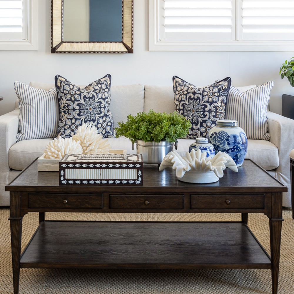 Huxley coffee table styled in front of  a neutral sofa with navy embroidered and navy and white striped cushions. The coffee table is styled with a wooden bone inlay box, a ruffled marble bowl, two small blue and white ginger jars, a faux greenery arrangement and a carved bone inlay tray with decorative coral.