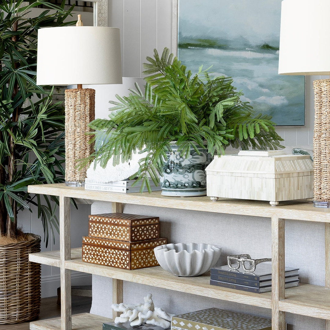 A light natural wood console table styled in a bright, modern living room with white paneled walls, two table lamps, potted green plants, and decorative baskets and boxes.