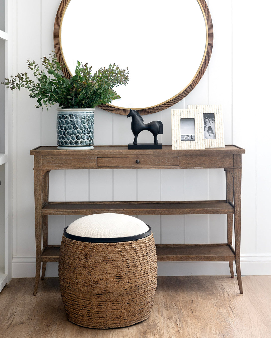 Wooden console table with round mirror, plant, and stool in a room.