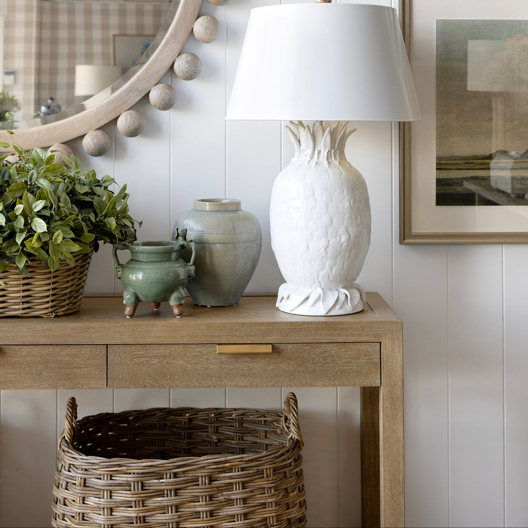 A white textured ceramic table lamp with a tapered shade sits on a light wood console table against a white shiplap wall, styled with a green plant, ceramic vases, a round beaded mirror, and framed landscape prints.