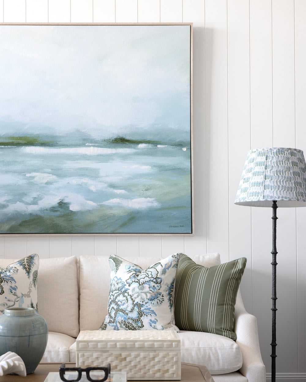 A large framed landscape painting of misty waves hangs on a white paneled wall above a cream sofa with patterned throw pillows. A wooden coffee table, a blue-gray ceramic vase, and a black floor lamp with a patterned shade are visible in the styled room.