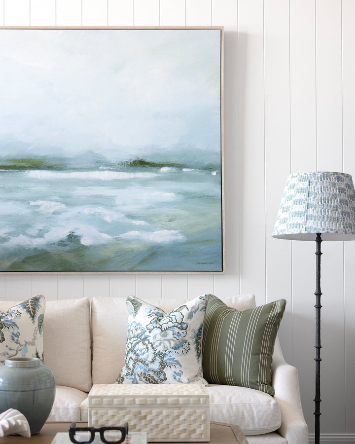 A large framed landscape painting of misty waves hangs on a white paneled wall above a cream sofa with patterned throw pillows. A wooden coffee table, a blue-gray ceramic vase, and a black floor lamp with a patterned shade are visible in the styled room.