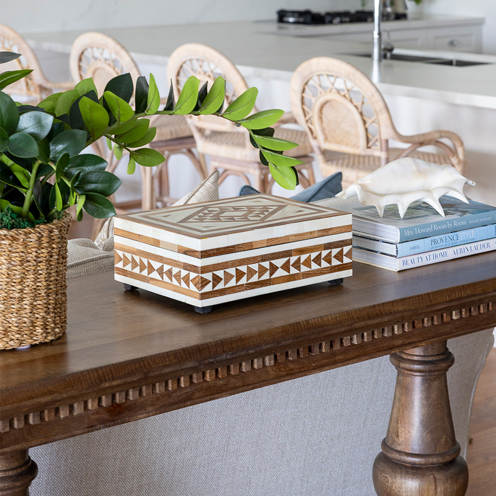 Laurent console styled with an artificial zanzibar plant in a rattan pot, a teak and bone inlay box and a stack of three books with a shell on top. The console sits with a sofa placed behind in a living room setting