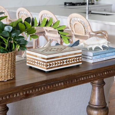 Laurent console styled with an artificial zanzibar plant in a rattan pot, a teak and bone inlay box and a stack of three books with a shell on top. The console sits with a sofa placed behind in a living room setting