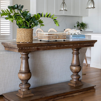 Laurent console styled with an artificial zanzibar plant in a rattan pot, a teak and bone inlay box and a stack of three books with a shell on top. The console sits with a sofa placed behind in a living room setting