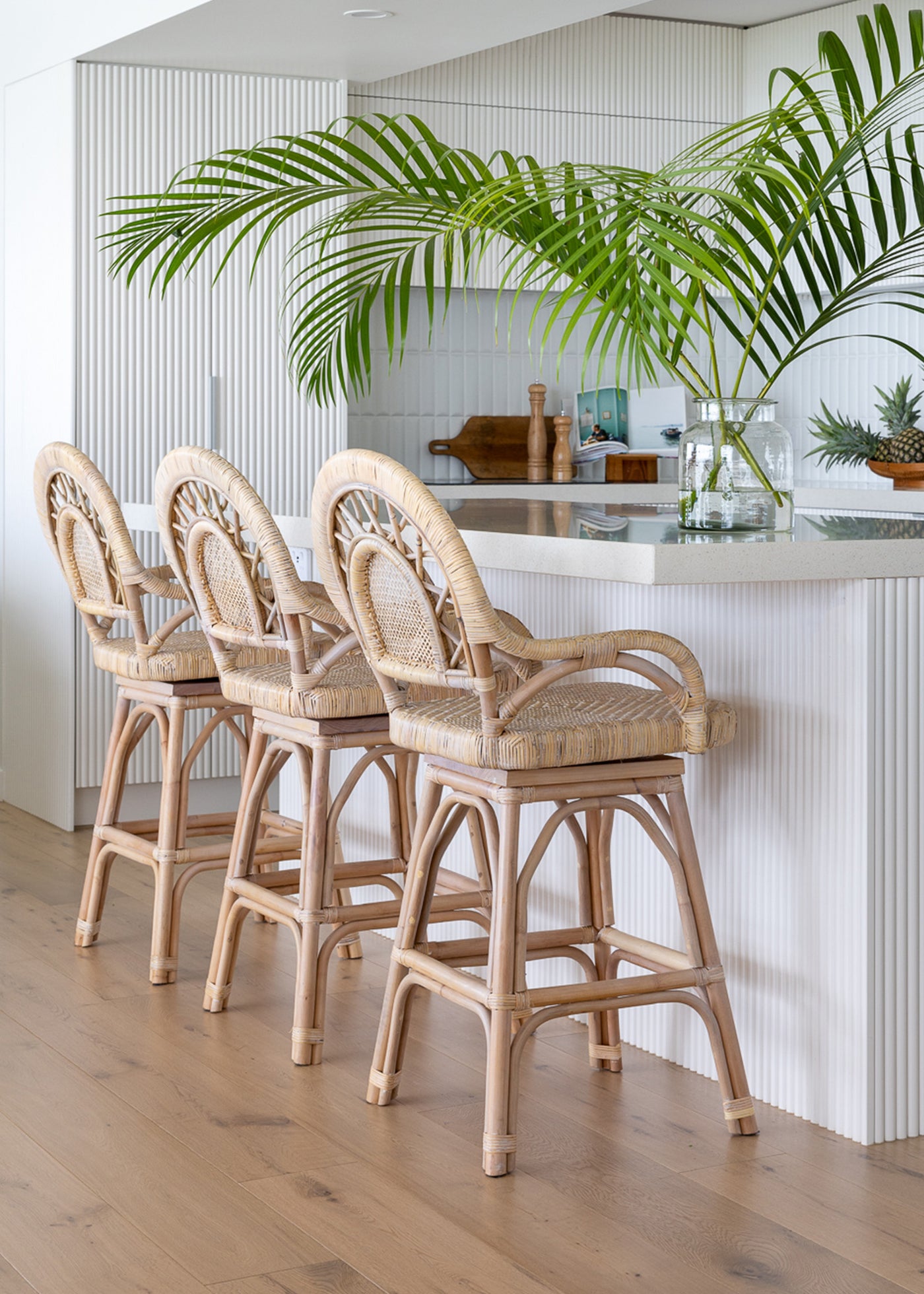 Styled image of 3 light lola bar stools at a kitchen counter, the kitchen is all white with a large clear vase of palm leaves