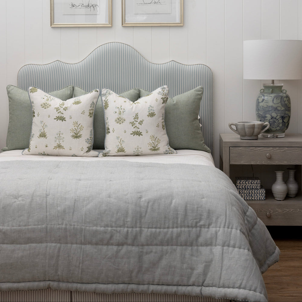 A styled bedroom scene featuring a bed with a light gray ribbed upholstered headboard, gray bedding, and two decorative pillows with a white botanical pattern. Above the bed are two framed botanical prints on a white shiplap wall.