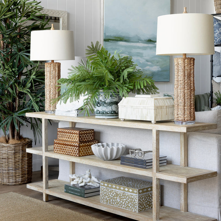 A light natural wood console table styled in a bright, modern living room with white paneled walls, two table lamps, potted green plants, and decorative baskets and boxes.