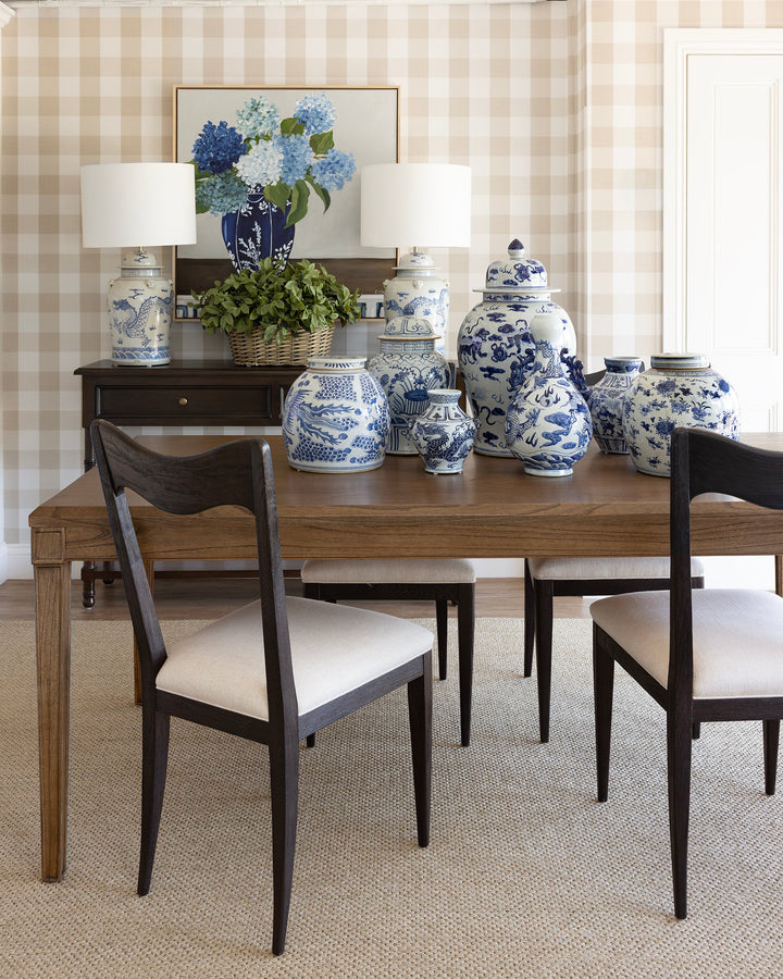 A styled dining room scene featuring a wooden dining table with four dark wood chairs that have cream upholstered seats, set against a beige gingham checkered wall. On the table are several blue and white porcelain ginger jars, and on the dark wood sideboard behind it are two matching blue and white table lamps and a framed floral print.
