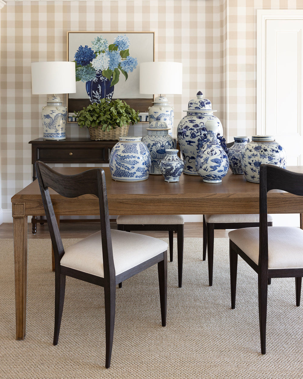 A styled dining room scene featuring a wooden dining table with four dark wood chairs that have cream upholstered seats, set against a beige gingham checkered wall. On the table are several blue and white porcelain ginger jars, and on the dark wood sideboard behind it are two matching blue and white table lamps and a framed floral print.