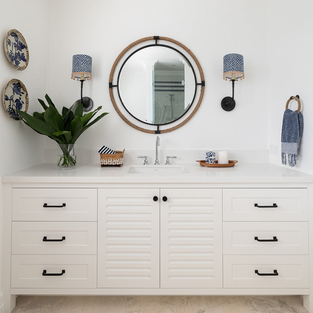 Round Melville Mirror sitting above a bathroom sink with one blue & white sconce either side. A vase of greenery on the bench top.