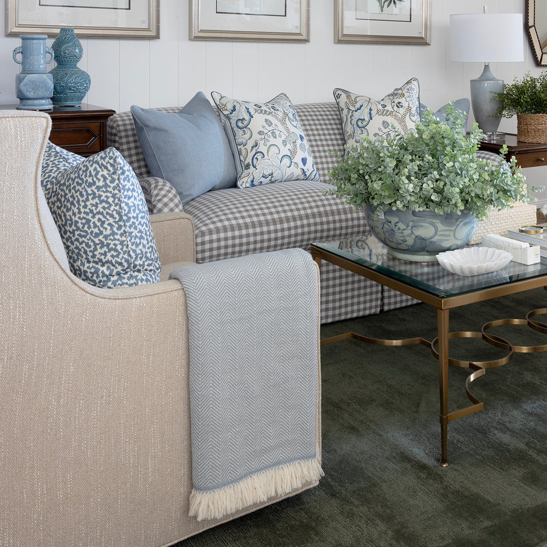 A styled living room interior featuring a beige armchair, a blue-and-white checkered sofa with blue accent pillows, a glass-top coffee table with gold legs, framed botanical prints on the wall, and a white ceiling with exposed beams.