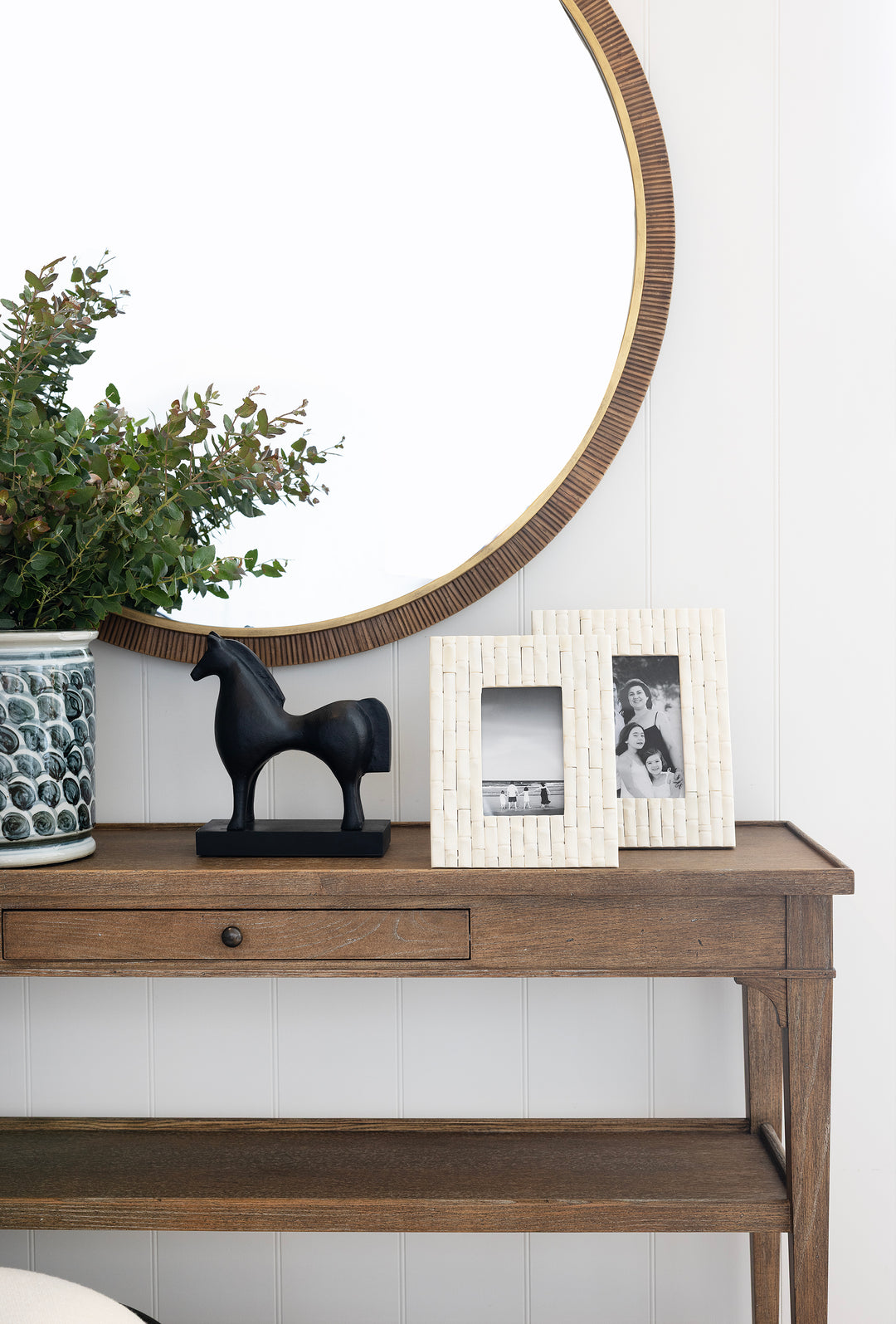 Wooden console table with decorative items including a horse statue, plant, and framed pictures against a white wall.