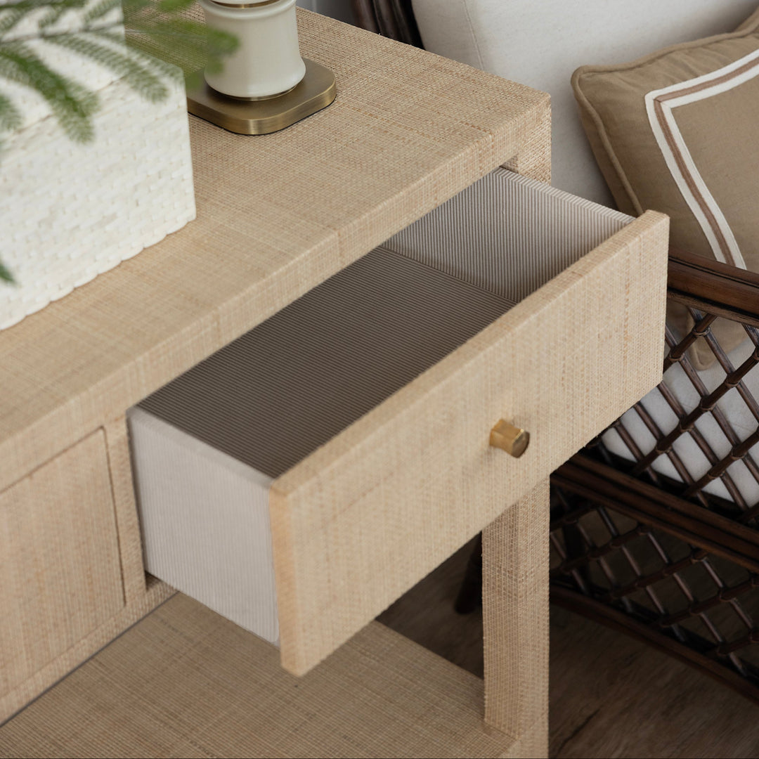 A close-up of a light beige woven wood nightstand with an open drawer showing a gray interior, styled next to a white table lamp and a cushioned chair.