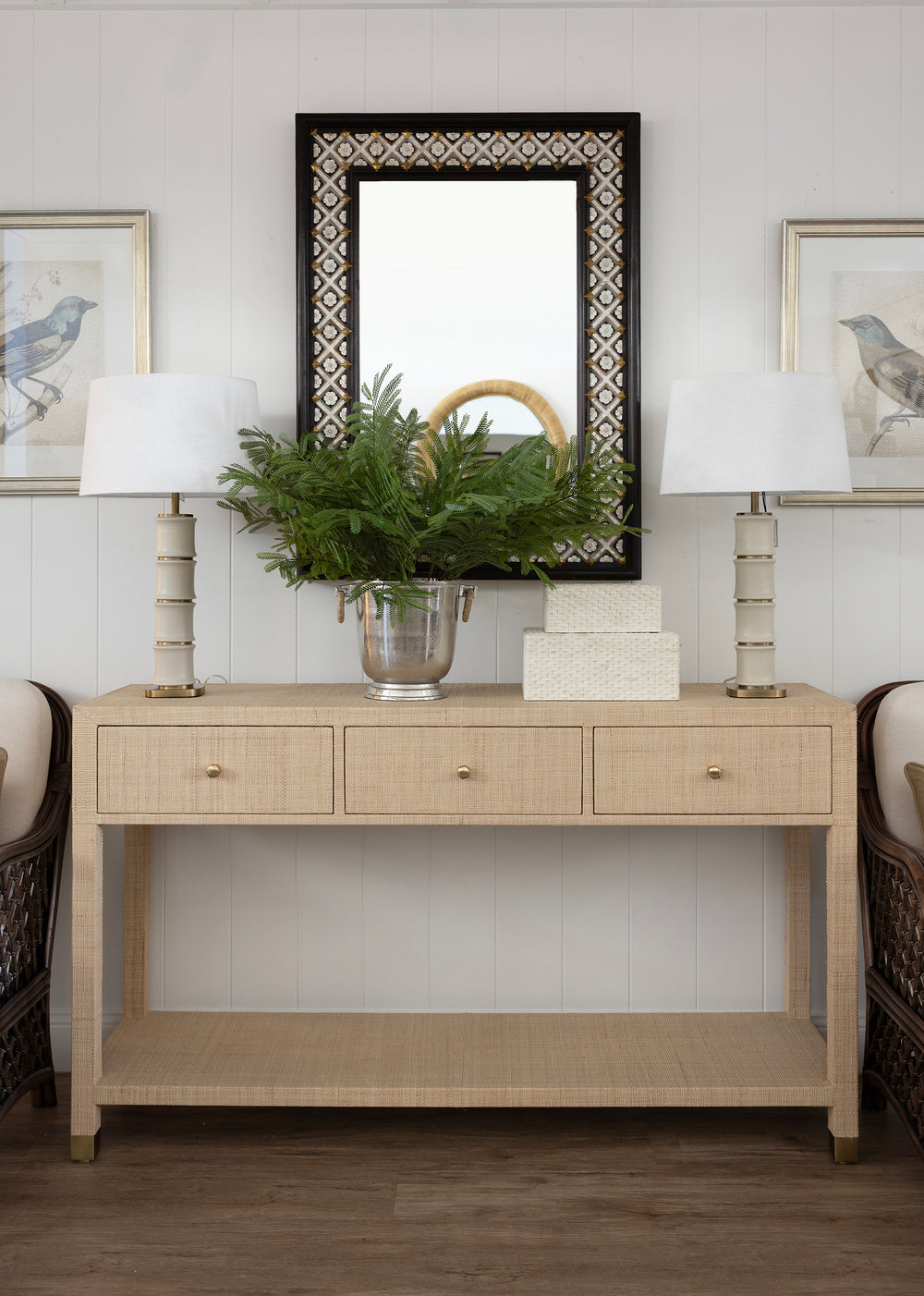 A light beige console table with three drawers and an open lower shelf, styled in a bright entryway with two white table lamps, a green fern in a silver vase, a white box, and a black-framed mirror above.