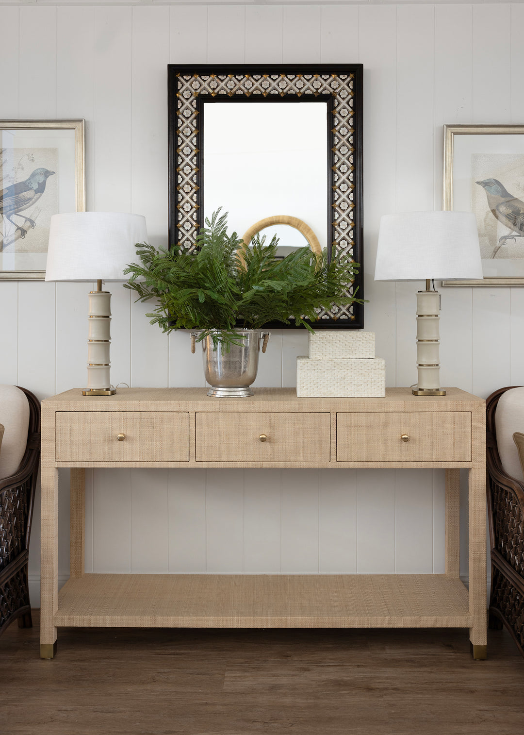 A light beige console table with three drawers and an open lower shelf, styled in a bright entryway with two white table lamps, a green fern in a silver vase, a white box, and a black-framed mirror above.