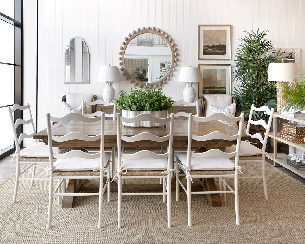 A dining room scene featuring a rustic wooden dining table surrounded by six white ladder-back chairs with white upholstered seats. The room is styled with a large round decorative mirror, white table lamps, framed art, potted green plants, and a white sideboard on a light beige rug.