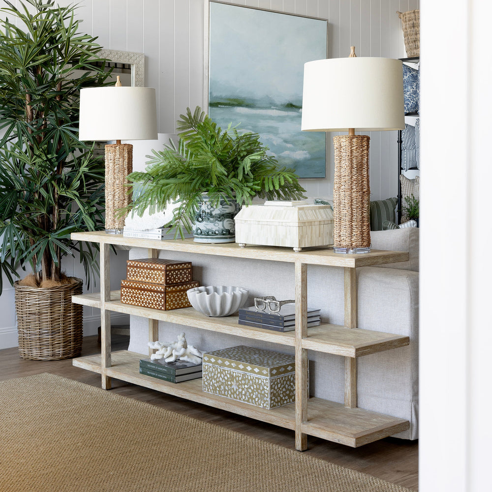 A light natural wood console table styled in a bright, modern living room with white paneled walls, two table lamps, potted green plants, and decorative baskets and boxes.