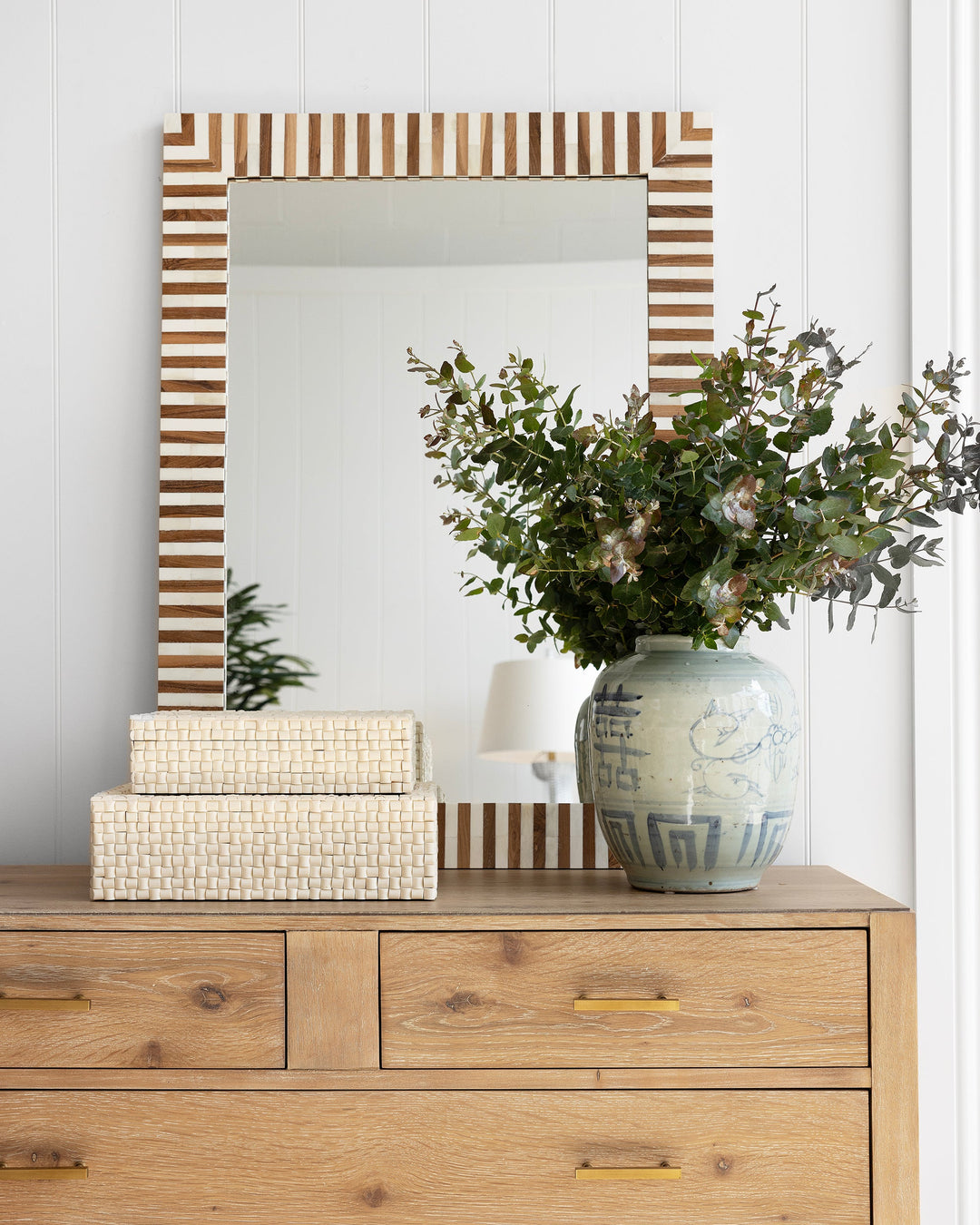 Wooden dresser with decorative items including a striped mirror, boxes, and a vase with flowers.