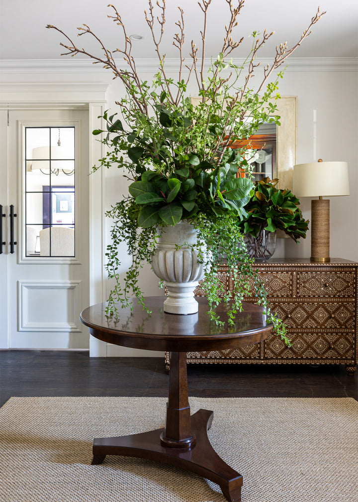 Thomas pedestal table in an entry way, styled on a sisal rug with a large faux greenery arrangement. Behind the table is a large bone inlay and teak chest with a lamp and fresh magnolia leaves in a vase.