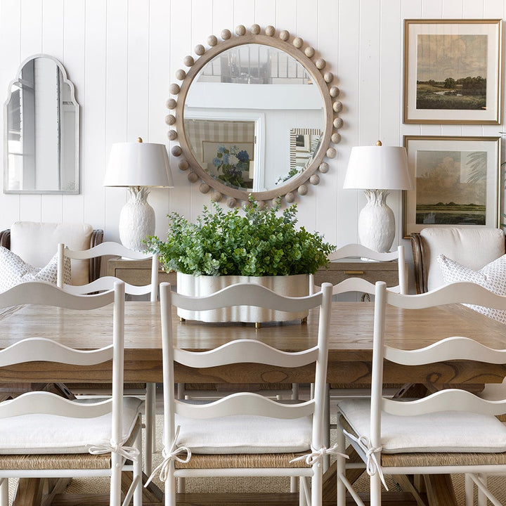 A dining room scene featuring a rustic wooden dining table surrounded by six white ladder-back chairs with white upholstered seats. The room is styled with a large round decorative mirror, white table lamps, framed art, potted green plants, and a white sideboard on a light beige rug.